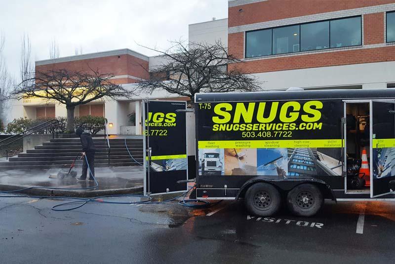 Snugs Services technician performing professional exterior building cleaning at a commercial property using specialized pressure washing equipment. by Snugs Services