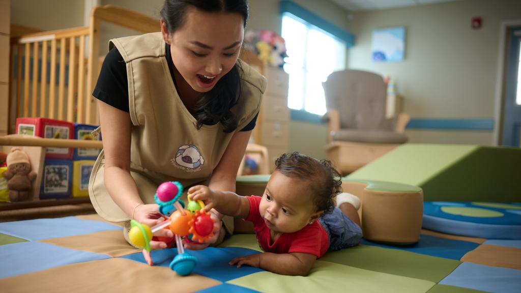 Primrose Schools infant teacher engaging baby with colorful toy during tummy time in early learning classroom by Primrose School at Vista Ridge