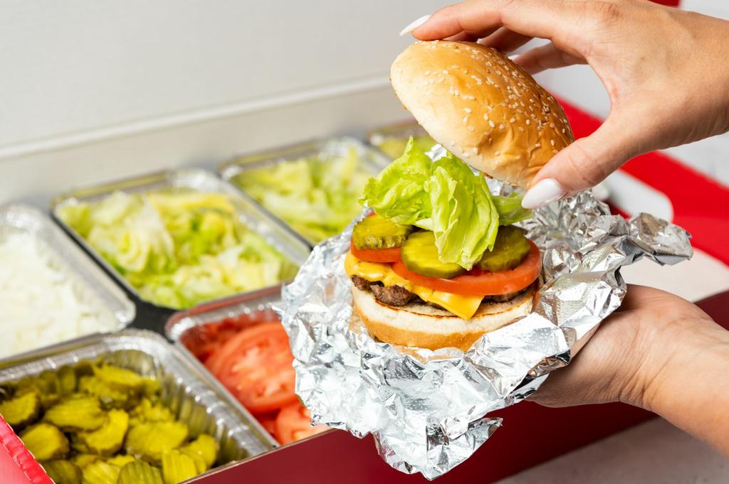 A close-up photo of a person putting the top bun of their cheeseburger in front of a Five Guys catering box of toppings. by Five Guys